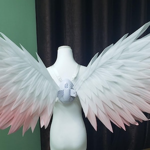 May include: A pair of large, expansive wings featuring a gradient color scheme that transitions from white at the top to a soft, pale pink at the tips. The wings are attached to a central white harness worn on a mannequin torso, displaying layered feather-like structures.