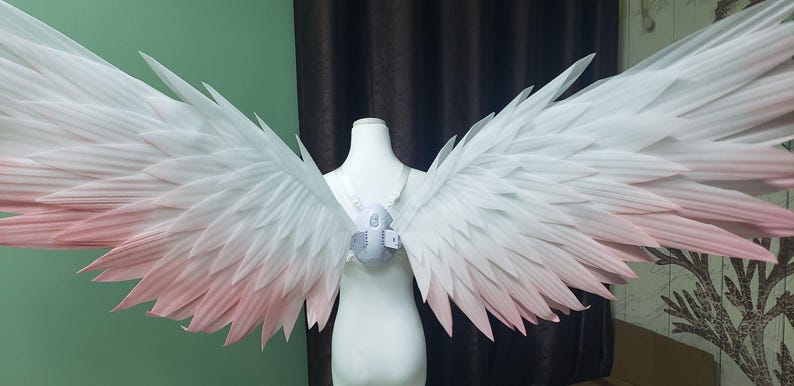 May include: A pair of large, expansive wings featuring a gradient color scheme that transitions from white at the top to a soft, pale pink at the tips. The wings are attached to a central white harness worn on a mannequin torso, displaying layered feather-like structures.