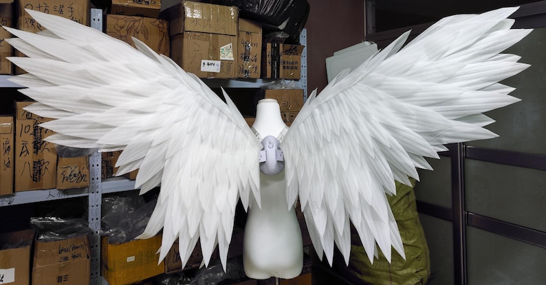 May include: A pair of large, white, layered feather-style wings attached to a central harness worn on a white mannequin torso. The wings feature pointed, overlapping segments that extend outward, creating a wide, expansive span of white plumage.