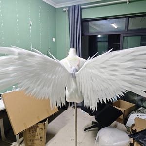 Puede incluir: Un par de alas blancas grandes y articuladas montadas sobre el torso de un maniquí. Las alas presentan múltiples capas de segmentos puntiagudos en forma de pluma que se extienden hacia afuera, unidas por un arnés central de color blanco.