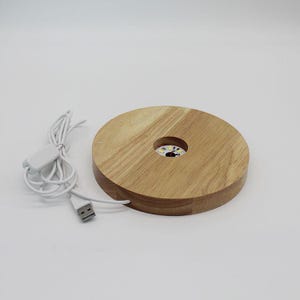 Op de afbeelding: Ronde houten LED-lichtbasis met een USB-kabel en aan/uit-schakelaar. De basis is gemaakt van lichtgekleurd hout en heeft een cirkelvormige uitsparing in het midden. De USB-kabel en schakelaar zijn wit.