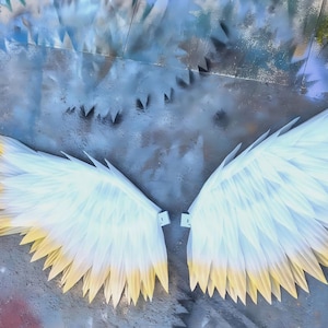 May include: A pair of large, stylized wings featuring layered, pointed feathers that transition from a crisp white at the base to a soft yellow at the tips. The wings are displayed flat, showing a symmetrical arrangement of individual feather segments.