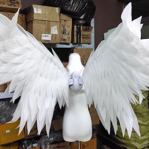 May include: A pair of large, white, feathered-style wings attached to a central harness worn on a white mannequin torso. The wings feature multiple layers of pointed, overlapping white segments that extend outward to create a wide, expansive span.