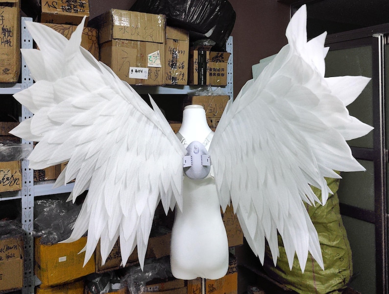 May include: A pair of large, white, feathered-style wings attached to a central harness worn on a white mannequin torso. The wings feature multiple layers of pointed, overlapping white segments that extend outward to create a wide, expansive span.