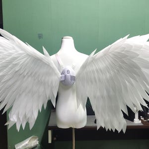 May include: A pair of large, white, articulated wings attached to a central harness worn on a mannequin torso. The wings feature numerous layered, pointed, feather-like segments extending outward, creating a wide, expansive wingspan design.