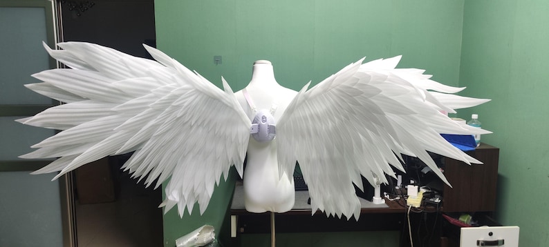 May include: A pair of large, white, articulated wings attached to a central harness worn on a mannequin torso. The wings feature numerous layered, pointed, feather-like segments extending outward, creating a wide, expansive wingspan design.