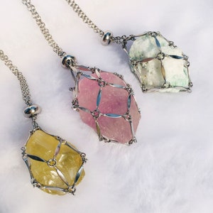 Könnte beinhalten: Drei silberne Halsketten mit Kristallanhängern, die in einem silbernen Käfig eingeschlossen sind. Die Kristalle sind rosa, grün und gelb.