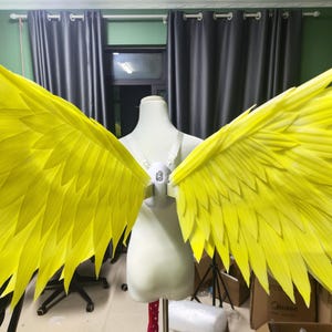 May include: Bright yellow costume wings with a feathered design, displayed on a mannequin. The wings are a vibrant shade of yellow and are spread wide, showcasing their full span. The wings are attached to a white harness.