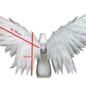 May include: Large white feathered wings attached to a central harness. The wingspan is marked with red lines indicating a width of 85-90 cm and a vertical height of 50 cm. The structure is mounted on a white torso mannequin form for display.