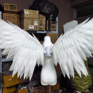 May include: A pair of large, white, articulated wings attached to a central harness worn on a mannequin torso. The wings feature multiple layers of pointed, feather-like segments extending outward in a wide, symmetrical span, creating a detailed avian appearance.