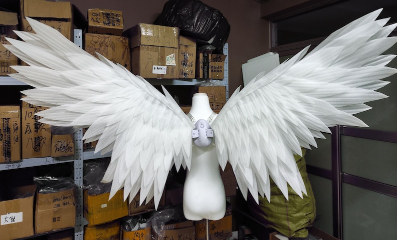 May include: A pair of large, white, articulated wings attached to a central harness worn on a mannequin torso. The wings feature multiple layers of pointed, feather-like segments extending outward in a wide, symmetrical span, creating a detailed avian appearance.