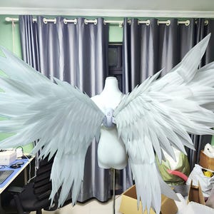 Puede incluir: Un par de alas de ángel blancas y articuladas montadas sobre un torso de maniquí blanco. Las alas presentan múltiples capas de segmentos puntiagudos en forma de pluma que se extienden hacia afuera en una amplia envergadura con aspecto detallado.