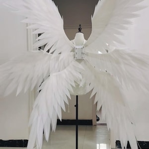 May include: A large set of white mechanical wings mounted on a mannequin torso. The wings feature multiple layers of pointed, feather-like panels extending outward from a central white rectangular motor housing worn on the back of the mannequin.