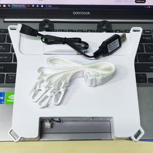 May include: A white rectangular plastic device featuring four corner slots, a black USB charging cable with a label reading USB Charger, and a white adjustable fabric strap with three white plastic swivel clips attached to the main unit.