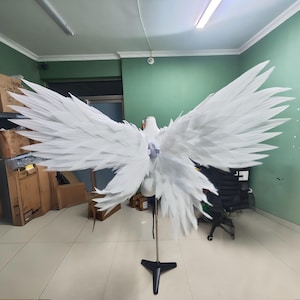 May include: Large, white angel wings with layered feathers, displayed on a black stand. The wings are spread wide, showcasing their full span. The background is a room with green walls and cardboard boxes.