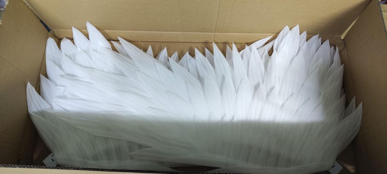 May include: A collection of white, pointed, feather-like foam pieces arranged in a layered, overlapping pattern. The individual elements are elongated, tapered, and have a smooth, matte texture, creating a dense, wing-like structure.