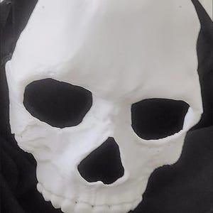 May include: A white skull mask with large eye sockets and a triangular nasal cavity. The mask has a smooth, matte finish and is set against a dark background. The mask is a Halloween or costume accessory.