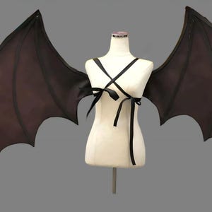 May include: Large dark brown bat wings attached to a white mannequin torso. The wings feature a structured frame with pointed tips and scalloped edges. Black ribbon straps cross over the chest and tie at the front to secure the wings in place.