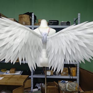 May include: Large white feathered wings attached to a central harness worn on a mannequin torso. The wings feature layered, pointed white feathers extending outwards in a wide span, secured by a white plastic harness with pink straps at the back.