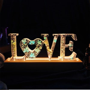 Può includere: Un cartello LOVE in acrilico trasparente con un cuore al centro. Le lettere sono riempite di fiori secchi e illuminate da dietro. Il cartello è su una base di legno.