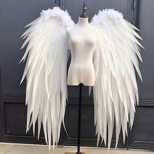 Puede incluir: Un par de alas de ángel grandes, blancas y puntiagudas unidas a un torso de maniquí blanco. Las alas presentan estructuras largas, delgadas y semirrígidas en forma de pluma que se extienden hacia abajo, con plumas suaves en los hombros.