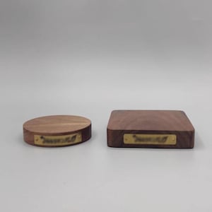 Puede incluir: Dos expositores de madera. Uno es un disco redondo y plano, y el otro es un bloque cuadrado. Ambos están hechos de madera oscura y tienen una pequeña placa de latón rectangular adjunta en la parte delantera.