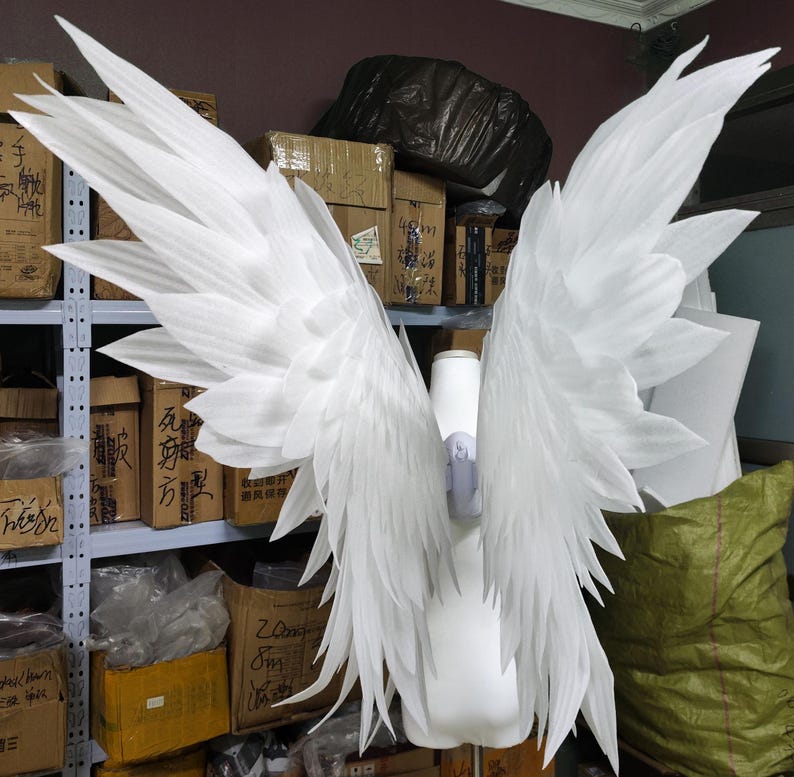 May include: A pair of large, white, feathered-style wings constructed from foam material. The wings feature multiple layers of pointed, overlapping segments that extend outward and upward, attached to a central white torso-shaped support structure.