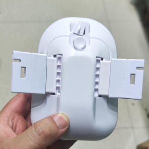 May include: A white plastic device featuring a rounded main body with a central circular button and a top sliding switch. Two rectangular, notched arms extend horizontally from the sides, each containing a series of small, evenly spaced square slots.