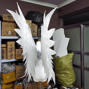May include: A pair of large, white, pointed feather-like wings attached to a central harness on a white mannequin torso. The wings feature multiple layers of elongated, sharp-edged white plumes that extend upward and outward in a symmetrical display.