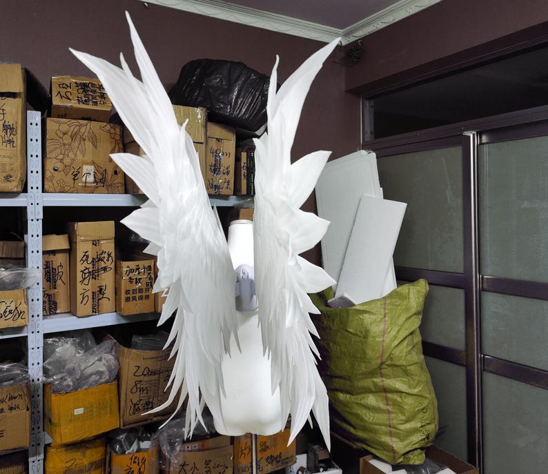 May include: A pair of large, white, pointed feather-like wings attached to a central harness on a white mannequin torso. The wings feature multiple layers of elongated, sharp-edged white plumes that extend upward and outward in a symmetrical display.