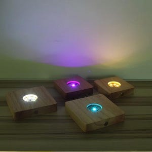 Pode incluir: Quatro bases de exibição LED quadradas de madeira com luzes de cores diferentes: branco, roxo, amarelo e azul. Cada base tem uma luz circular no centro e um pequeno botão na lateral. As bases são feitas de madeira clara.