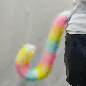 May include: A rainbow-colored faux fur tail, featuring a gradient of pink, yellow, blue, and white. The tail is attached to a person wearing a white top and black skirt. The tail is fluffy and curved.