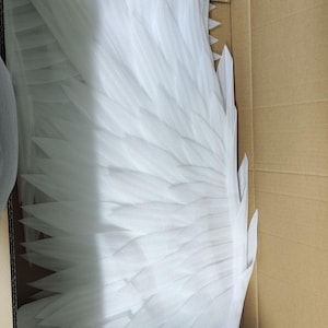 May include: A pair of large, white, layered foam wings with pointed, feather-like segments. The wings are packed inside a brown cardboard box, showing a structured, symmetrical design with multiple overlapping sections that create a wing shape.