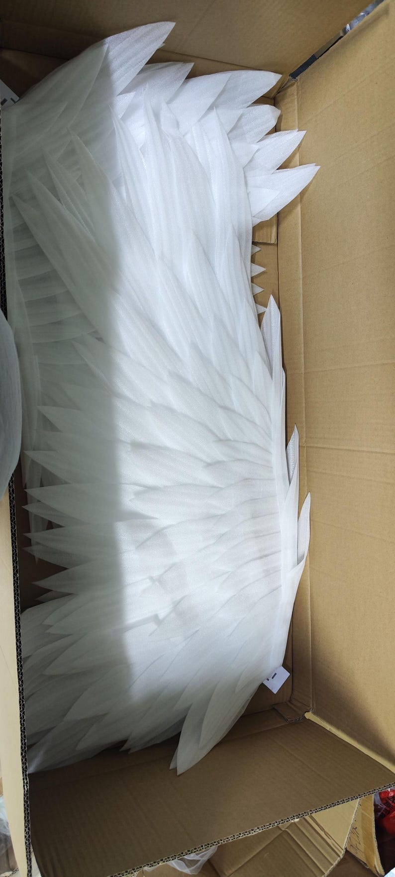 May include: A pair of large, white, layered foam wings with pointed, feather-like segments. The wings are packed inside a brown cardboard box, showing a structured, symmetrical design with multiple overlapping sections that create a wing shape.