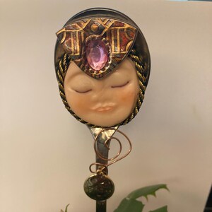 Op de afbeelding: Een decoratieve lepel met een gezichtsontwerp. De lepel heeft een gezicht met gesloten ogen, een roze edelsteen en een goud-bronzen hoofdtooi. De lepel is bevestigd aan een zwarte steel met een groene bol en koperdraaddetails.