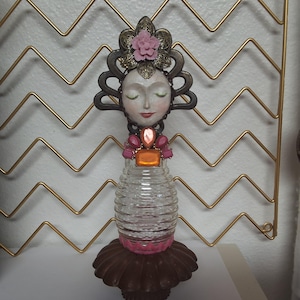 May include: A decorative figurine featuring a serene face with closed eyes, adorned with a floral headdress and jewel accents. The figure's body is a clear, ribbed glass bottle, resting on a brown, ruffled base. The overall height is approximately 8 inches.