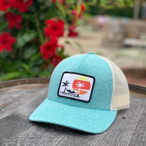 以下が含まれることがあります： 白のメッシュバックが付いたターコイズブルーのトラッカーハット。ハットの前方には、ヤシの木と「No Wrong Place」という文字が書かれたパッチが付いています。