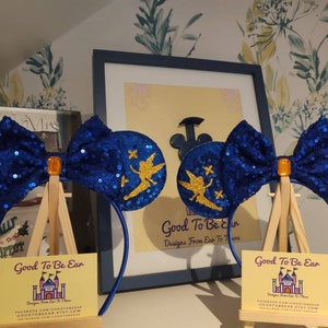 May include: Blue and gold Tinkerbell mouse ears with a sparkly blue bow. The ears are on a headband and are displayed on a small easel. The text on the easel reads "Good To Be Ear Designs From Ear To There".