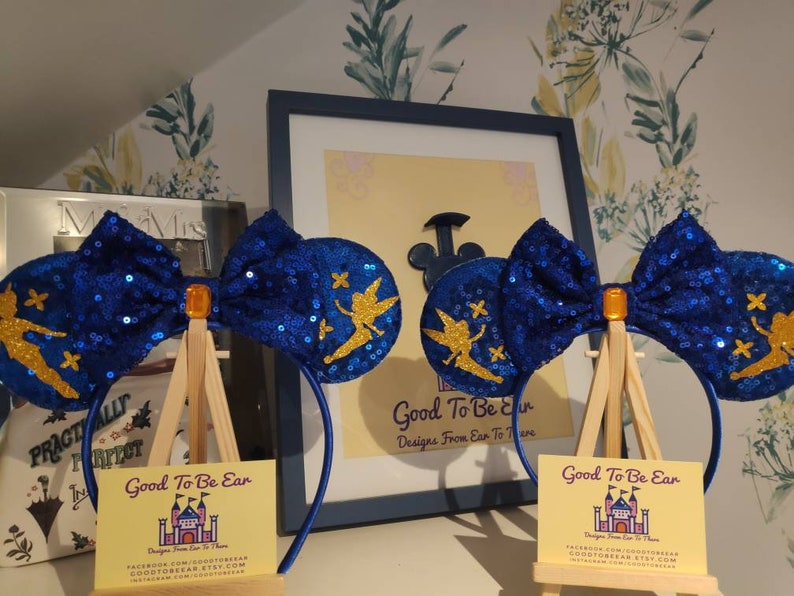 May include: Blue and gold Tinkerbell mouse ears with a sparkly blue bow. The ears are on a headband and are displayed on a small easel. The text on the easel reads "Good To Be Ear Designs From Ear To There".