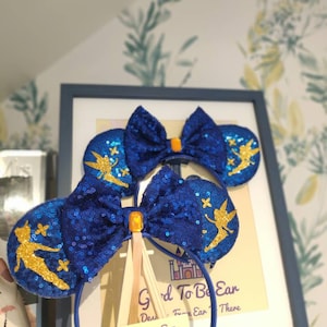 May include: Blue and gold Tinkerbell mouse ears with a blue bow and a gold glitter finish. The ears are on a headband with a tag that says "Good To Be Ear" and a castle graphic.