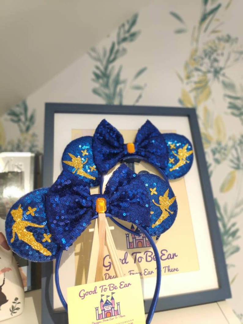 May include: Blue and gold Tinkerbell mouse ears with a blue bow and a gold glitter finish. The ears are on a headband with a tag that says "Good To Be Ear" and a castle graphic.