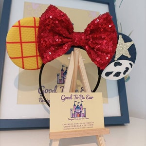 Op de afbeelding: Een rood pailletten strik hoofdband met een geel en rood geruit oor aan de linkerkant en een blauw en wit koe-print oor aan de rechterkant. De hoofdband wordt getoond op een kleine houten ezel met een visitekaartje waarop staat "Good To Be Ear Designs From Ear To There" en contactgegevens voor Facebook, Etsy en Instagram.