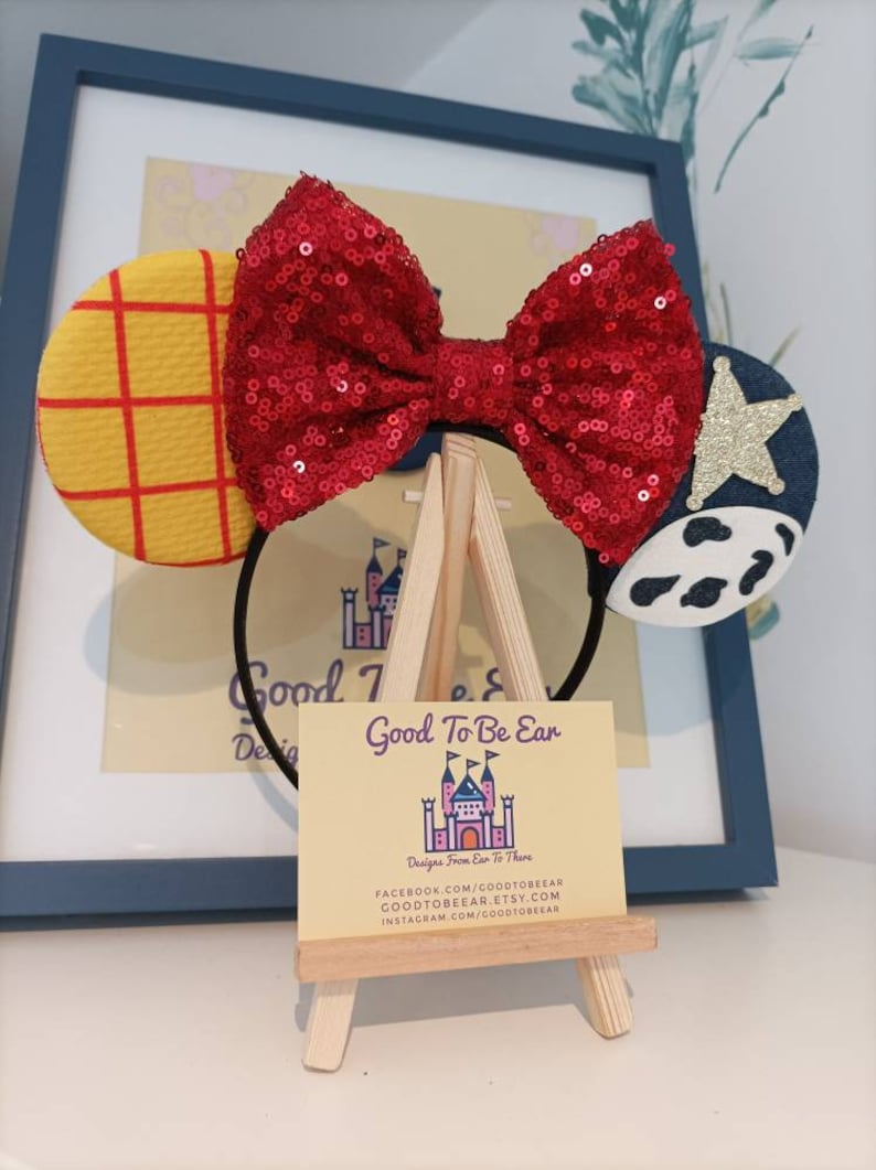 Op de afbeelding: Een rood pailletten strik hoofdband met een geel en rood geruit oor aan de linkerkant en een blauw en wit koe-print oor aan de rechterkant. De hoofdband wordt getoond op een kleine houten ezel met een visitekaartje waarop staat "Good To Be Ear Designs From Ear To There" en contactgegevens voor Facebook, Etsy en Instagram.
