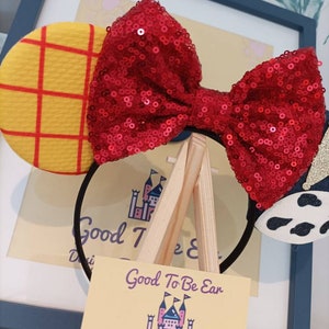 Op de afbeelding: Een rood haarband met een strik van pailletten, een geel en rood geruit oor en een zwart-witte koe-print oor. De haarband wordt getoond op een houten ezel met een visitekaartje waarop staat "Good To Be Ear, Designs From Ear To There, FACEBOOK.COM/GOODTOBEEAR, GOODTOBEEAR.ETSY.COM, INSTAGRAM.COM/GOODTOBEEAR".