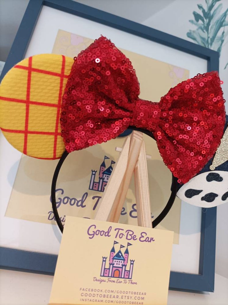 Op de afbeelding: Een rood haarband met een strik van pailletten, een geel en rood geruit oor en een zwart-witte koe-print oor. De haarband wordt getoond op een houten ezel met een visitekaartje waarop staat "Good To Be Ear, Designs From Ear To There, FACEBOOK.COM/GOODTOBEEAR, GOODTOBEEAR.ETSY.COM, INSTAGRAM.COM/GOODTOBEEAR".