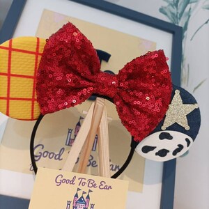 Op de afbeelding: Een rood pailletten strik hoofdband met een geel en rood geruit oor aan de ene kant en een blauw oor met een witte koe print en een gouden ster aan de andere kant. Het hoofdband wordt getoond op een kleine houten ezel met een visitekaartje waarop staat "Good To Be Ear Designs From Ear To There" met contactgegevens eronder.