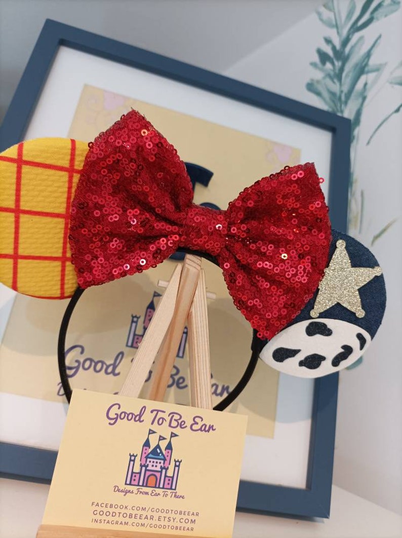 Op de afbeelding: Een rood pailletten strik hoofdband met een geel en rood geruit oor aan de ene kant en een blauw oor met een witte koe print en een gouden ster aan de andere kant. Het hoofdband wordt getoond op een kleine houten ezel met een visitekaartje waarop staat "Good To Be Ear Designs From Ear To There" met contactgegevens eronder.