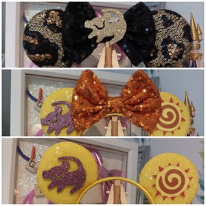 May include: A set of three pairs of handmade Disney-themed Minnie Mouse ears. The first pair features a black and gold bow with a gold lion silhouette. The second pair features an orange bow with a purple lion silhouette. The third pair features a yellow bow with a sun design.