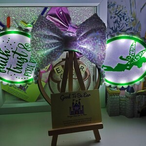 May include: A sequined bow with a purple centre is displayed on a small wooden easel. Two illuminated circular signs with green borders and text are on either side. A miniature castle is in the background.