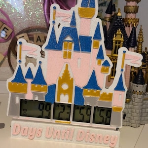 May include: A white, pink, blue and gold 3D printed countdown clock shaped like Cinderella's castle. The clock displays "Days Until Disney" and the numbers 09 23 59 59.